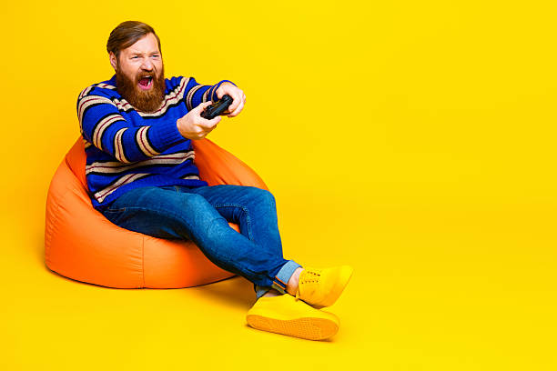 istock Young bearded man sitting on an orange beanbag chair holding a controller, expressing joy against a vibrant yellow background 2235748028