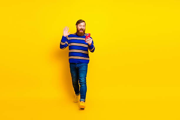 istock Cheerful red-haired man with beard waving hand while holding smartphone against vibrant yellow background 2235748096