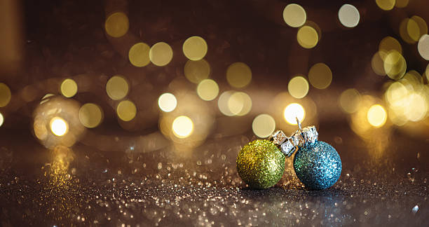Close-up of two glitter Christmas baubles in green and blue on a sparkling surface with warm golden bokeh lights in the background. Elegant festive decoration symbolizing celebration, winter holidays, and seasonal joy.