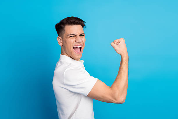 istock Young confident man posing against blue background, flexing arm and expressing joy and energy in casual white shirt. 2235750899