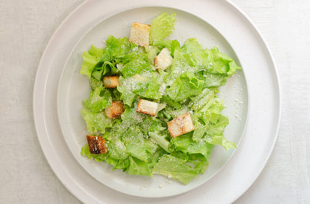 Traditional Caesar salad with croutons and parmesan cheese in a ceramic flat plate with ingredients on a gray table. Traditional American cuisine concept. Vertical orientation. Selective focus. Top view.