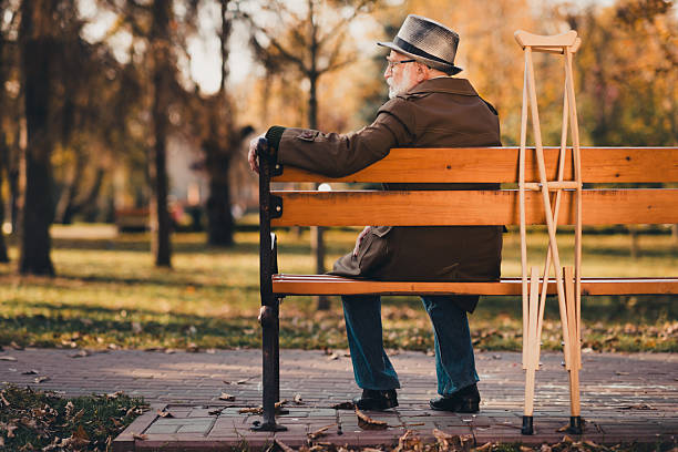 A senior man wearing a hat and coat sitting on a wooden bench in an autumn park. Crutches are near, symbolizing resilience and solace amid nature's beauty during a peaceful seasonal day.