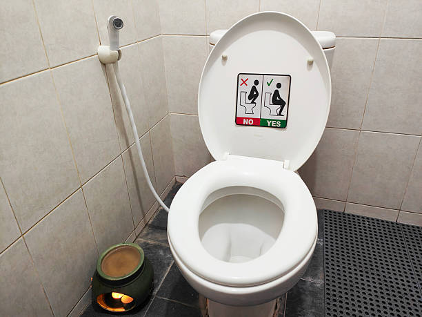 close-up shot of a white toilet with an instruction sticker on the lid showing correct and incorrect sitting positions, placed inside a tiled bathroom with a bidet spray and small floor candle.\n\nConcept: toilet, restroom, bathroom interior, toilet seat, hygiene, toilet instruction sign, bidet spray, sanitary, toilet etiquette, toilet usage guide, wc interior