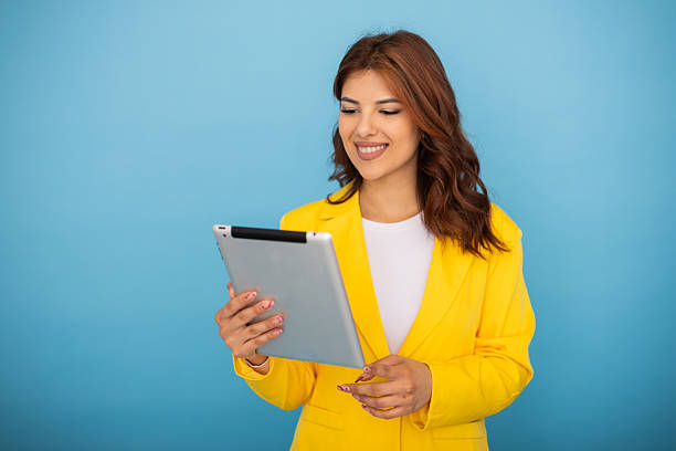 istock Confident professional woman working on tablet, wearing bright yellow jacket against blue studio backdrop, expressing positive workplace attitude 2235758328
