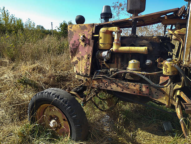 An old, rusty tractor stands in tall dry grass, partially disassembled, with traces of rust and peeling paint. Left to its own devices, it symbolizes a past of agricultural use, contrasting with its current neglect and oblivion.