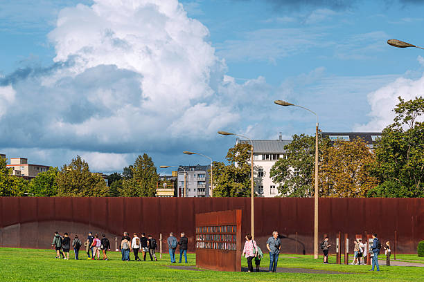 A diverse group of people visit the steel pillars of the Berlin Wall Memorial, a historical landmark surrounded by green grass and urban buildings, symbolizing reconciliation and remembrance in modern Berlin.
