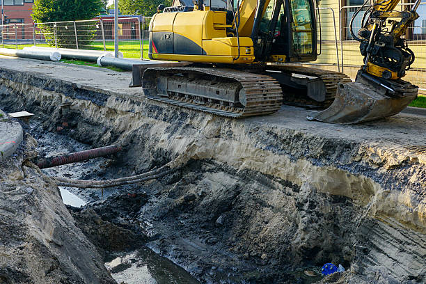 A yellow excavator stands beside a deep trench exposing pipes and soil layers, part of construction and infrastructure repair work