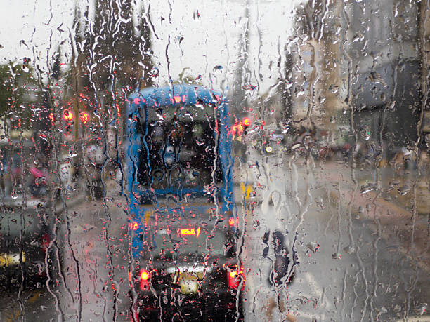 A city bus driving down a wet street with rain falling. The bus is partially obscured by the raindrops hitting the windshield, giving a sense of movement and atmospheric perspective. The sky is overcast, adding to the rainy day ambiance. Raindrops on windshields create patterns and streaks that add visual interest