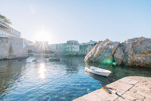 White rowboat tied to stone pier in calm harbor water near ancient fortress walls at sunrise. Scenic coastal landscape with clear water and rocky shoreline. Peaceful maritime setting.