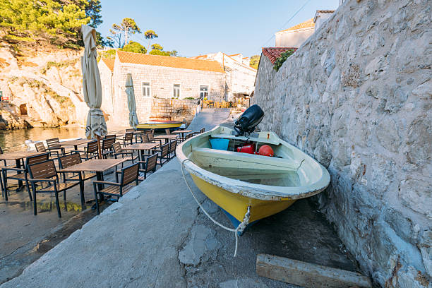 Small yellow motorboat tied up beside stone wall near empty outdoor cafe tables and umbrellas. Quiet coastal scene with Mediterranean architecture and calm waters. Seaside dining and boating setting.