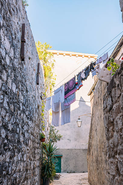 Laundry drying on clotheslines strung between stone buildings in a narrow alleyway. Rustic urban scene with textured walls and potted plants. Charming old town street atmosphere.