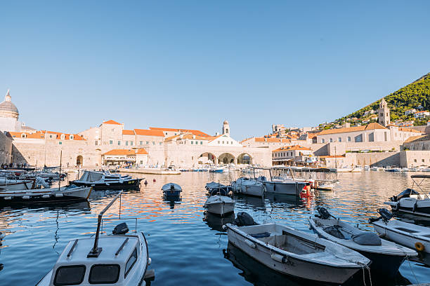 Small boats moored in a calm harbor with historic stone buildings and red rooftops in the background. Scenic coastal town with clear blue sky and peaceful waterfront. Mediterranean harbor view.