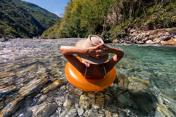 Adult woman wearing sun hat relaxing on orange float in crystal clear mountain river. Person enjoying calm water and nature in peaceful outdoor setting. Leisure and summer vacation concept.