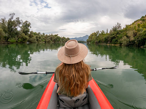 Young woman paddling a red kayak on a calm river surrounded by green trees. Outdoor adventure with peaceful water and cloudy sky. Solo kayaking in nature.