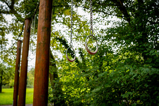Two gymnastic rings suspended on chains as part of an outdoor workout station. The installation is located in a park surrounded by trees and green leaves.