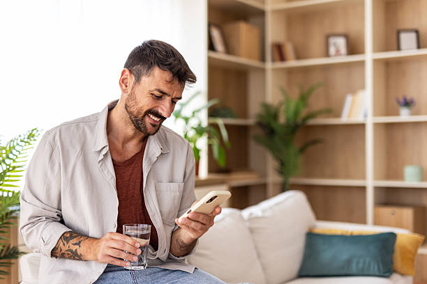 istock Smiling man sitting on sofa holding glass of water and using smartphone 2235798721