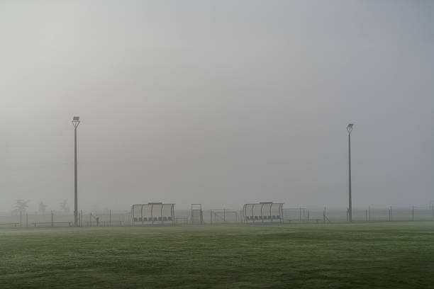 Empty sports field with benches and tall lights shrouded in morning fog.