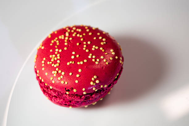 A plate of colorful macarons is displayed against a warm, textured background. These delicate confections come in various shades, including vivid red and light beige, with chocolate or cream fillings visible. Round in shape, one macaron is sprinkled with small yellow embellishments, adding texture and visual interest. The plate is white with subtle floral detailing, contributing to a cozy and inviting ambiance.