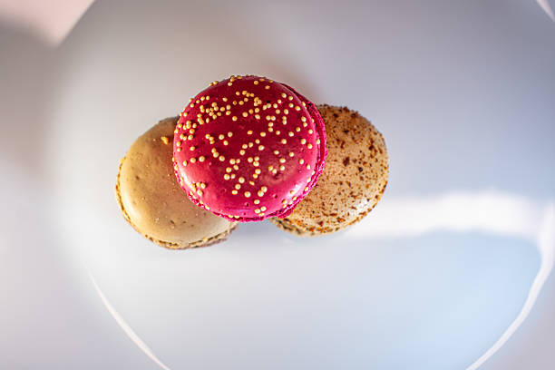 A plate of colorful macarons is displayed against a warm, textured background. These delicate confections come in various shades, including vivid red and light beige, with chocolate or cream fillings visible. Round in shape, one macaron is sprinkled with small yellow embellishments, adding texture and visual interest. The plate is white with subtle floral detailing, contributing to a cozy and inviting ambiance.