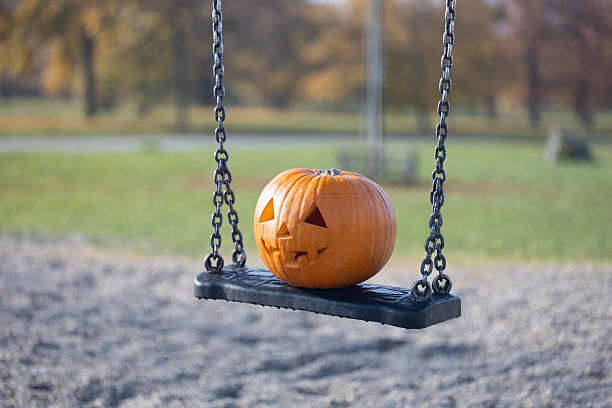 istock Jack-o'-lantern pumpkin with carved face resting on a swing in a playground, surrounded by autumn foliage and soft sunlight, creating a festive seasonal atmosphere 2235817698