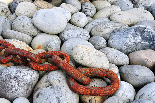 Santa Cruz, Flores Island, Azores, Portugal: close-up view of smooth, rounded grey stones of varying sizes, scattered across the ground, resting on top of the stones is a thick, rusted metal chain, its reddish-brown corrosion contrasting sharply with the pale, weathered surface of the rocks. The composition emphasizes texture and color, highlighting the interplay between the natural erosion of the stones and the decay of the man-made chain. Cobbles and chain.