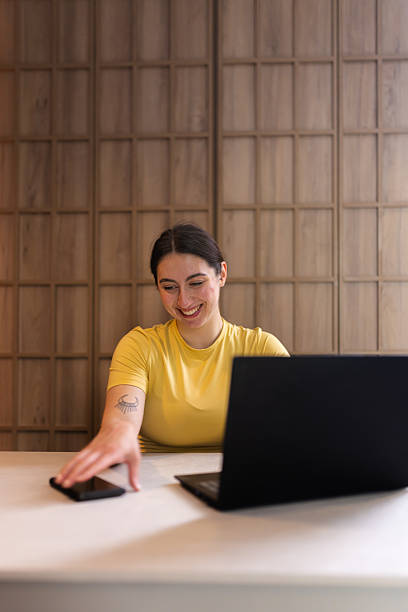 istock Young woman smiling while using a laptop and reaching for her phone in a cozy indoor workspace during the afternoon 2235827521