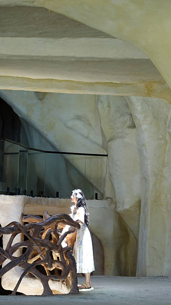 In the pictures, a scene under a coastal rock formation is presented. A woman is wearing a light-colored dress with pale purple patterns, brown sandals, and has a purple flower accessory in her long hair, along with sunglasses. She sometimes looks up, shading her eyes with her hand to gaze into the distance, and sometimes leans against the rock while holding a drink to enjoy the scenery. Around her are strangely shaped brown rocks. In the distance, there is the azure sea with islands and reefs, and the sky is clear. The whole scene is full of the freshness and comfort of a summer seaside, fully showing a leisurely vacation atmosphere.