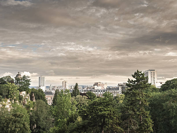 Elevated panorama toward Paris from Parc des Buttes Chaumont, showing a broad skyline with towers and rooftops above trees under a cloudy sky, conveying an aerial style city panorama.