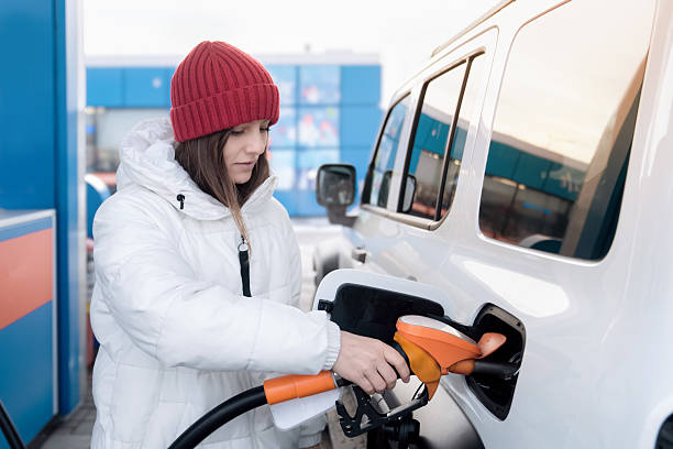 Expensive diesel and gasoline at the gas station. A woman stands at a gas station, carefully refueling her vehicle with gasoline. Dressed in a warm jacket and a red beanie, she holds the fuel nozzle stock photo