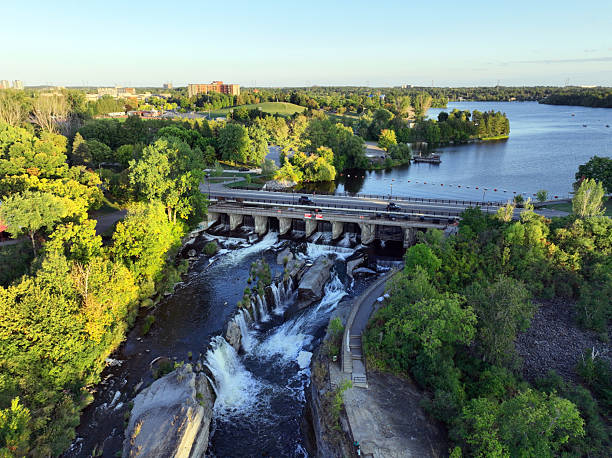 A river flowing past a small waterfall along with a hydro-electric dam and bridge, surrounded by lush green trees and a city skyline in the distance