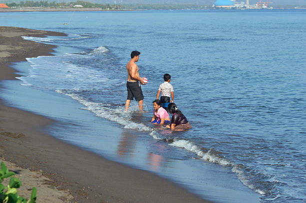 A father holds a ball and heads into shallow seawater as his son follows closely, while mother and daughter sit near the foamy shoreline.