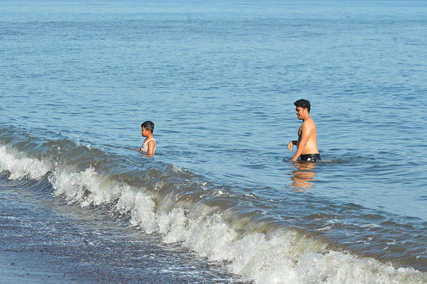 A father stands with his young son in shallow seawater at the beach, both enjoying a peaceful moment together in the gentle morning surf.