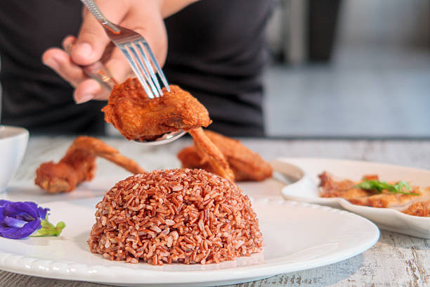 This close-up shot captures a delicious home-cooked meal featuring crispy fried chicken wings and nutritious brown rice. The scene is set on a light wooden table, with more chicken wings visible in the background and a vibrant purple butterfly pea flower adding a touch of color to the foreground. It evokes a sense of comfort and a wholesome dining experience.