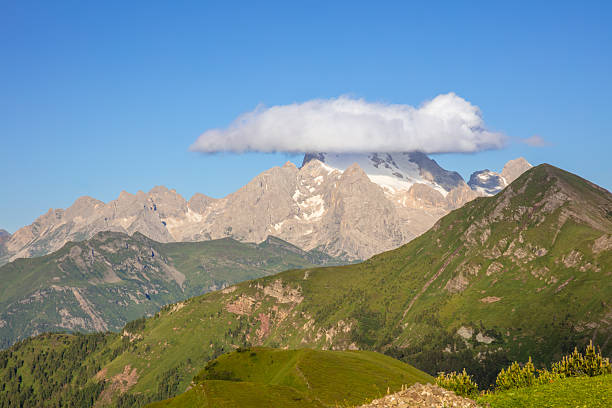 Sunny summer day in the Italian Dolomites. Green and rocky peaks. A lone cloud in the blue sky clings to a snowy peak