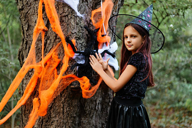 istock Young girl in witch costume decorating a tree for Halloween celebration in a forest setting during autumn season 2235859275