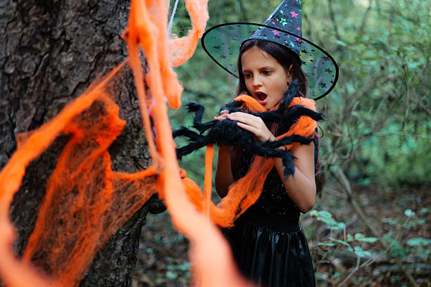 istock Young girl in witch costume interacting with spider decorations in a forest during Halloween preparations 2235859276