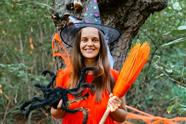 istock A cheerful witch in a vibrant red dress holds a broom and a plush spider while surrounded by Halloween decorations in a wooded area 2235859355