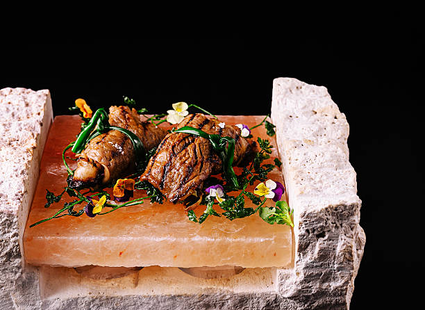 Grilled beef rolls tied with greens, served on Himalayan salt stone, decorated with edible flowers and herbs, fine dining presentation.