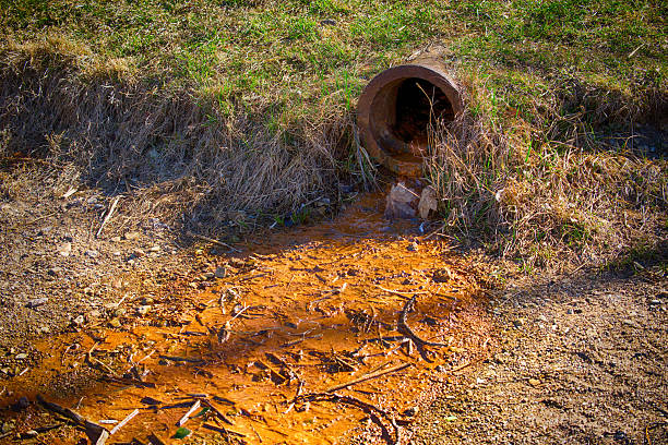 A rusty pipe discharges waste water into a small stream, creating an orange, polluted flow onto the ground.
