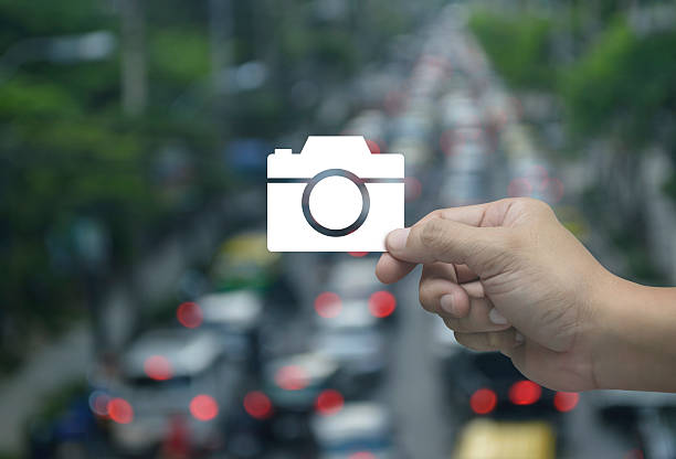 Hand holding camera flat icon in fingers over blur of rush hour road in city, Business camera service shop online concept