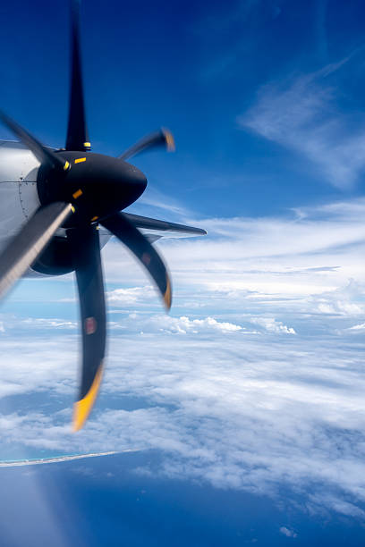 View from an airplane flying over the Pacific Ocean, with a spinning propeller in the foreground. The sky is partly cloudy, and the deep blue water extends to the horizon