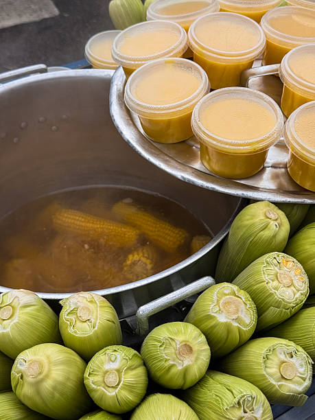 A vibrant display of fresh raw corn next to a large pot cooking flavorful broth. Containers of freshly portioned corn broth stacked beside them showcase the preparation process, reflecting traditional culinary art. The market atmosphere surrounds this detailed and vivid scene.