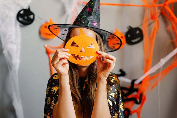 istock A person wears a spooky witch hat and holds a bright pumpkin mask while surrounded by vibrant Halloween decorations and cobwebs 2235883884