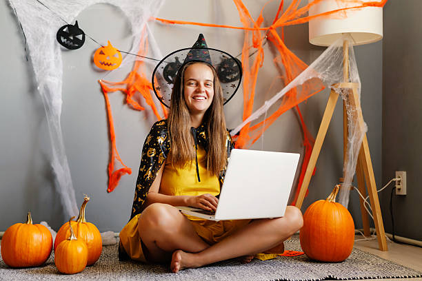 istock A girl wears a playful witch costume, smiling while using her laptop surrounded by pumpkins and Halloween decorations in a cozy space 2235883896