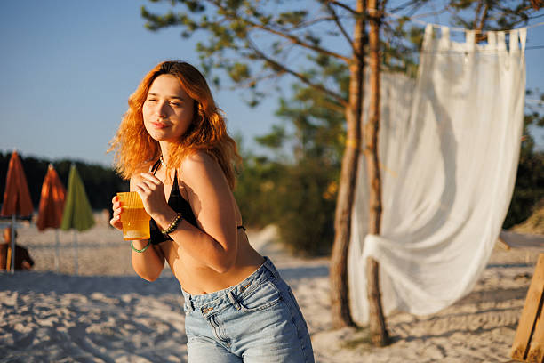 A vibrant young woman relaxes on a pristine beach, holding a frosty glass of tropical drink as sunlight glints off the condensation