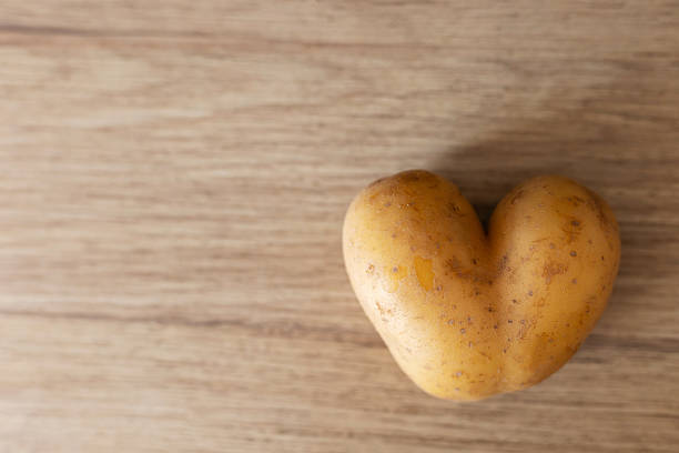 A unique heart-shaped potato rests on a smooth wooden table, showcasing its distinct shape and texture. The simple setting adds warmth, making it an intriguing sight for food lovers.