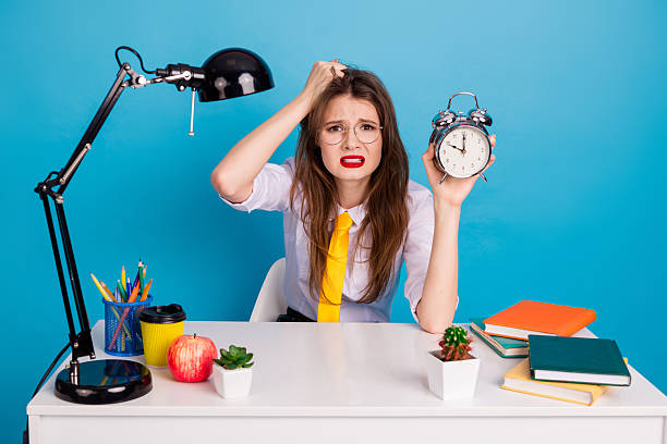 istock Stressed female student in vintage uniform holding an alarm clock while sitting at a desk with school supplies 2235897821