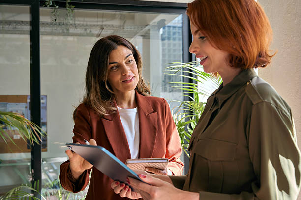 istock Two happy smiling business women talking using tab standing in green office. 2235900912