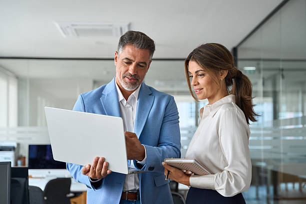 Busy professional smiling businessman and businesswoman leaders having conversation at work. Executives team of two business people working talking using laptop technology standing in corporate office