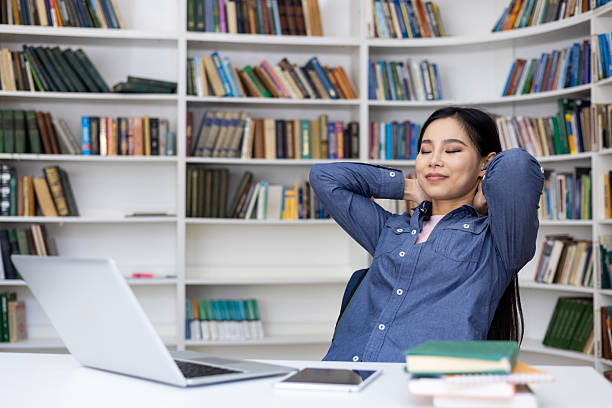 A relaxed Asian woman sits in a library with a laptop and books, resting with her hands behind her head.